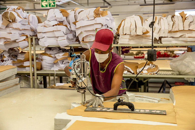 Young person in cap, facemask and work gloves handles a machine on a workbench, with piles of fabric stacked on shelves in the background