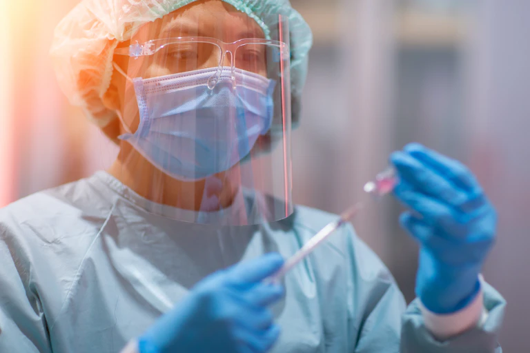 A doctor in full PPE holds a vaccine vial and syringe.