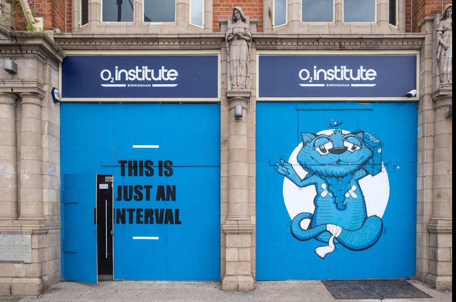 O2 Institute building with the words 'this is just an interval' painted in black against a blue door and on the door to the right, a painting of a bandaged blue cat carrying a boombox next to its head