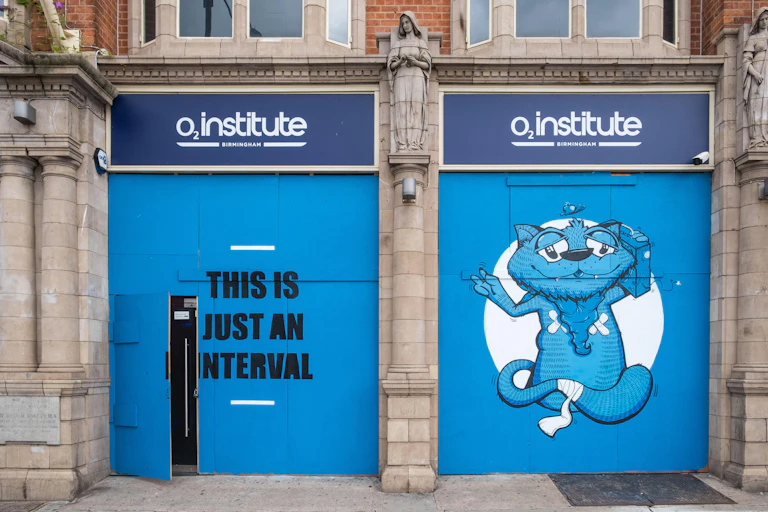 O2 Institute building with the words 'this is just an interval' painted in black against a blue door and on the door to the right, a painting of a bandaged blue cat carrying a boombox next to its head