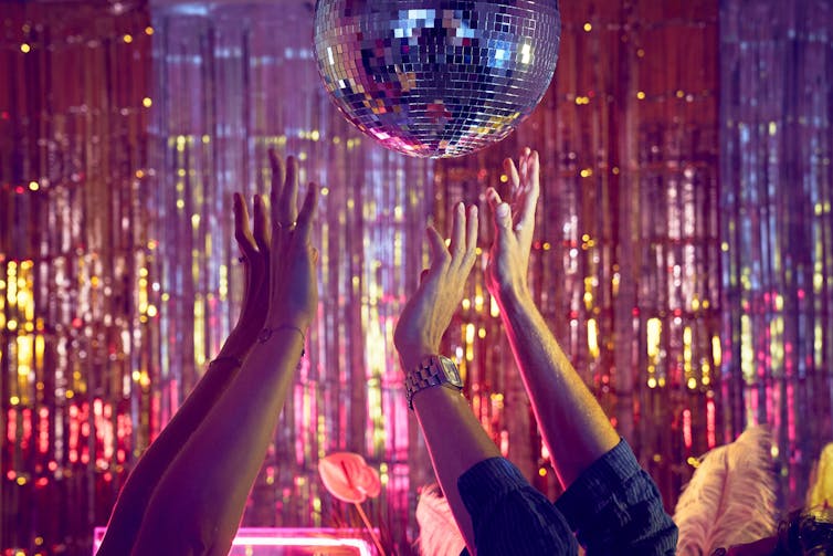Hands in the air in a club reach upwards towards a discoball in front of a metallic fringe curtain