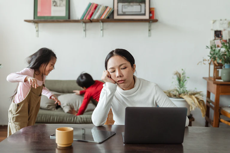 Mother sits on a laptop trying to get work done while children in the background playing