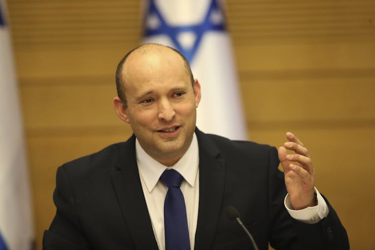 A man smiles and gestures in front of an Israeli flag