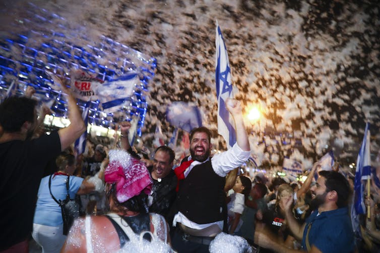 People smile and celebrate in a crowd, waving Israeli flags