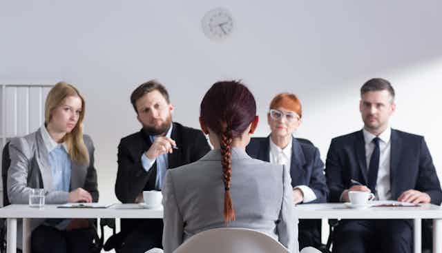 A woman is seen from behind sitting on a chair facing a panel of four other people during an interview.