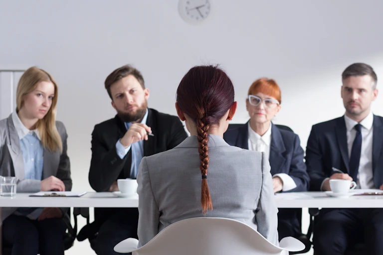 A woman is seen from behind sitting on a chair facing a panel of four other people during an interview.