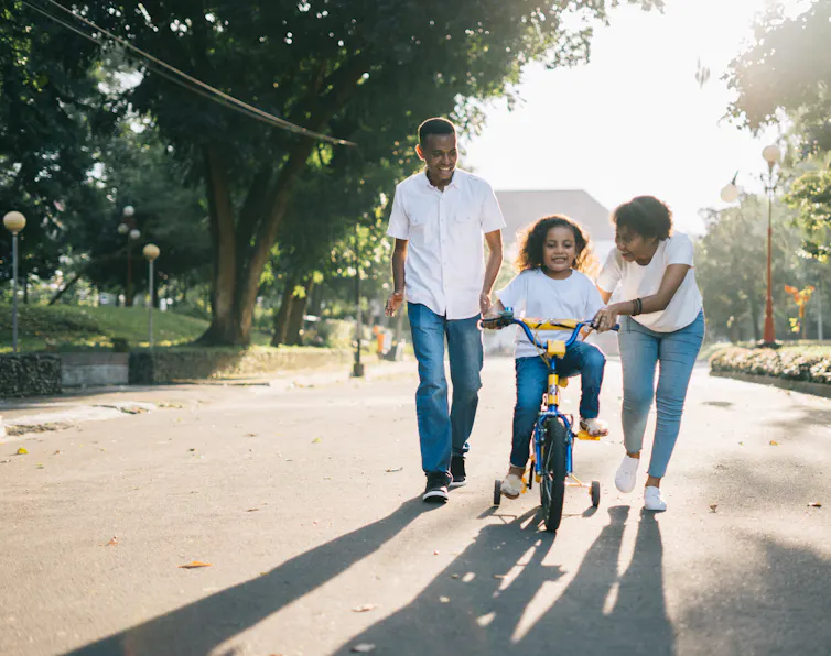 Family of three, mother and father push child on bike with training wheels down tree lined street.