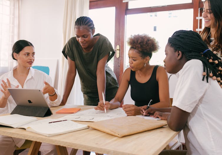 Five women at a desk have a conversation. 