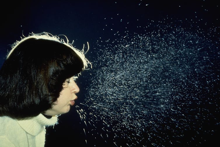 Person sneezing with visible droplets against black backdrop.