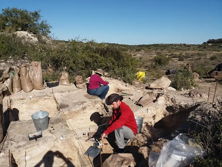 Two people, one in a red shirt and a brown hat, the other in a tan hat and a pink shirt, are using spades in a small cordoned off area against a backdrop of trees and hills.