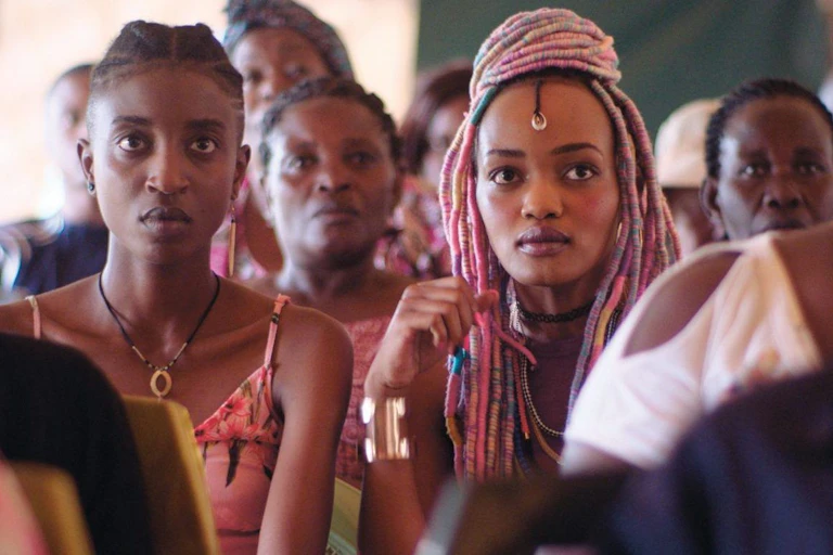 Pople sit in rows in what looks to be a church, looking ahead. The frame is dominated by two striking young women, one with pink braids.
