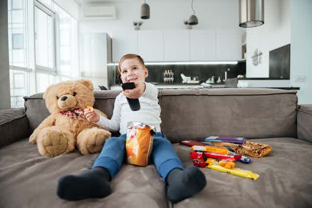Niño en sofá, con patatas fritas y otras chucherías, un osito y un mando a distancia.
