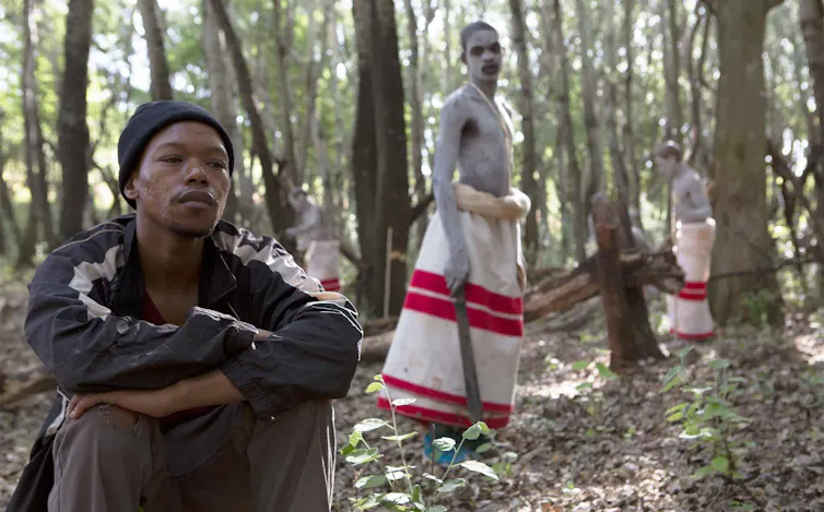 A young man sits, contemplative in Western clothing in a forest; behind him a young man in traditional clothing with white body paint looks at him.