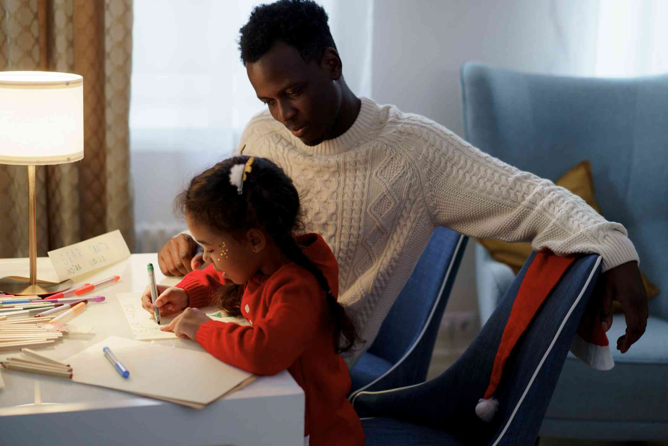 A father sits wtih daughter who is writing at a desk.