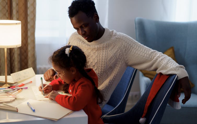 A father sits wtih daughter who is writing at a desk.