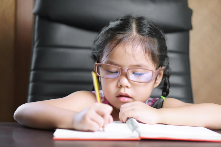 Young girl wearing glasses writes in a notebook.