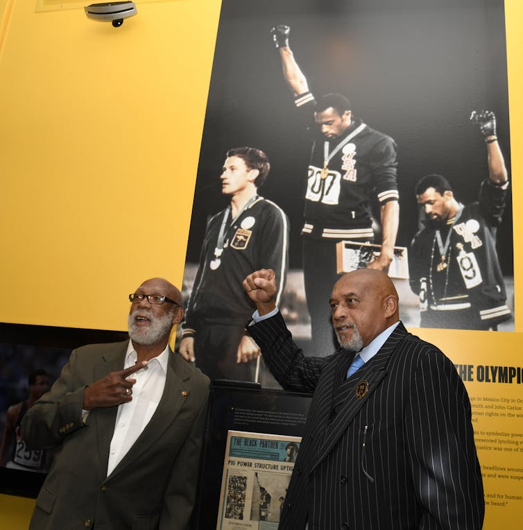 John Carlos and Tommie Smith stand in front of a photo of them raising Black Power salutes during the 1968 olympics