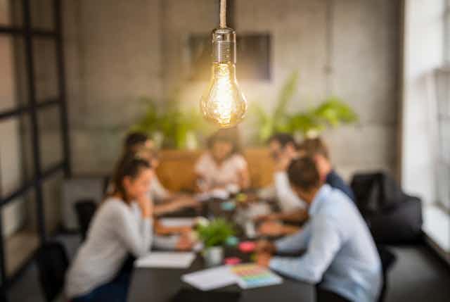 A lightbulb is in sharp focus over a meeting of young people in an office setting.