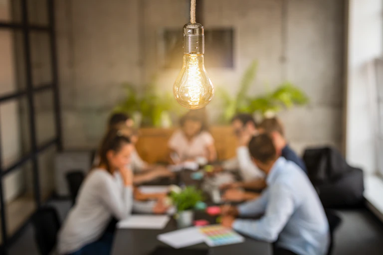 A lightbulb is in sharp focus over a meeting of young people in an office setting.