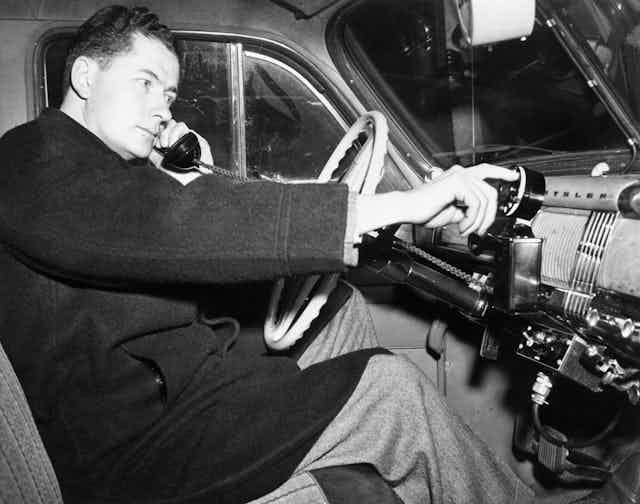 A historic photograph of a man sitting behind the wheel of a 1940s automobile holding telephone handset and dialing a telephone rotary dial installed in the car