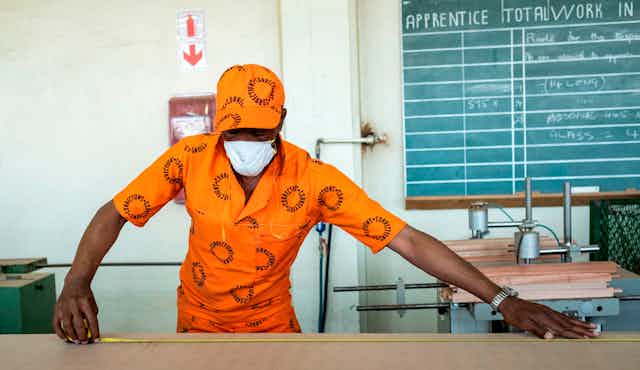 A man in an orange workout marked with the words 'correctional services' and a mask on his face is busy measuring a piece of wood, piles of cut wood and a blackboard behind him.