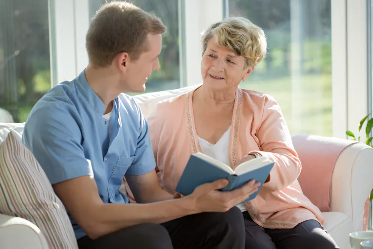 A young man reads a book with an elderly woman.