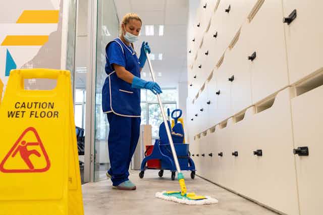 Woman in blue cleaning uniform and face mask sweeps the floor in front of caution wet floor sign