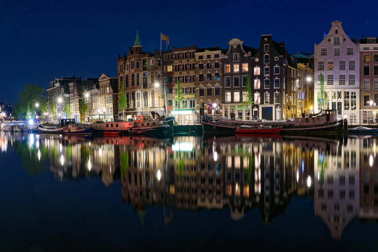 Buildings lit up against a night sky are reflected in the still waters of an Amsterdam canal