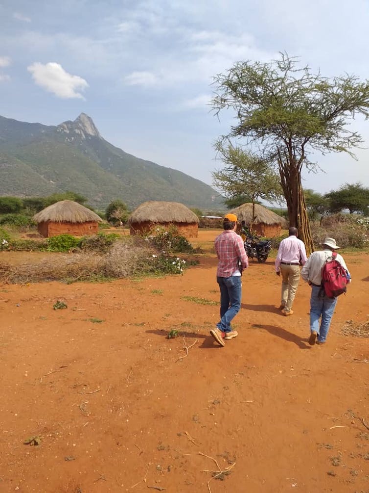 People walk across a reddish dirt area with grass-roofed huts in the background.