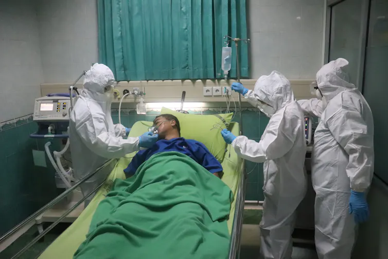 Man lies in green bed surrounded by three people in white protective suits.