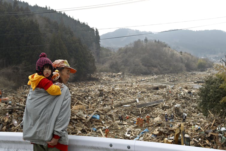 A small girl on her grandfather's back in front of a scene of devastation and rubble.