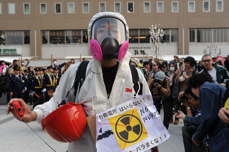 A person wearing a gas mask and a anti-nuclear sign stands in front of a crowd.