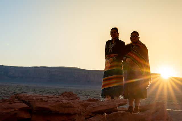 Two Navajo sisters dressed in traditional clothing standing in the desert with the sun behind them. 