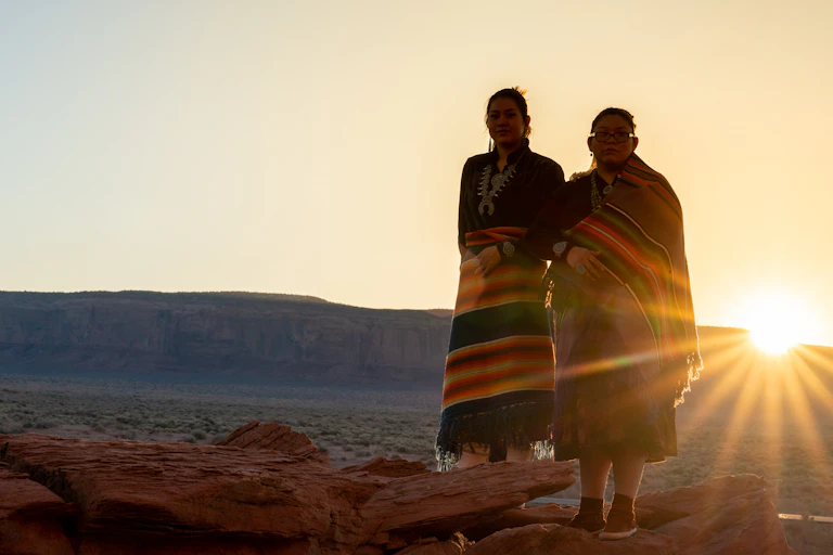 Two Navajo sisters dressed in traditional clothing standing in the desert with the sun behind them.