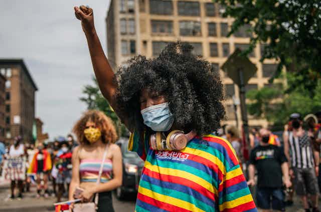 A Black girl wearing a rainbow flag raises her right fist in the middle of a crowded street.
