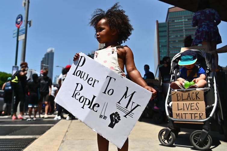 A little Black girl on a street holds a sign that reads 'Power to my people!!'