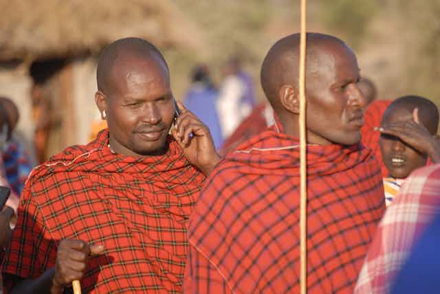 Two men in traditional Maasai dress; one holds a mobile phone to his ear.