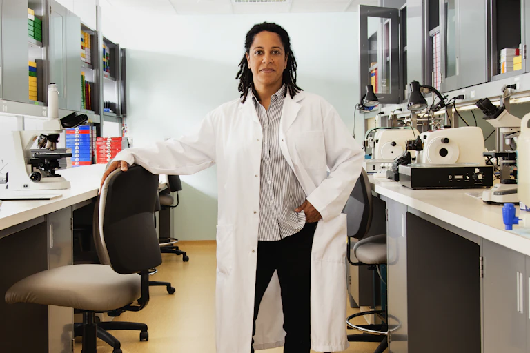 A woman in a white lab coat stands in the middle of a laboratory, her left hand in her pocket and her right hand on a chair