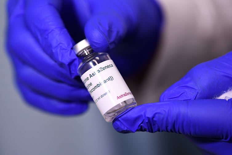 Gloved hands holding a vial of AstraZeneca vaccine.