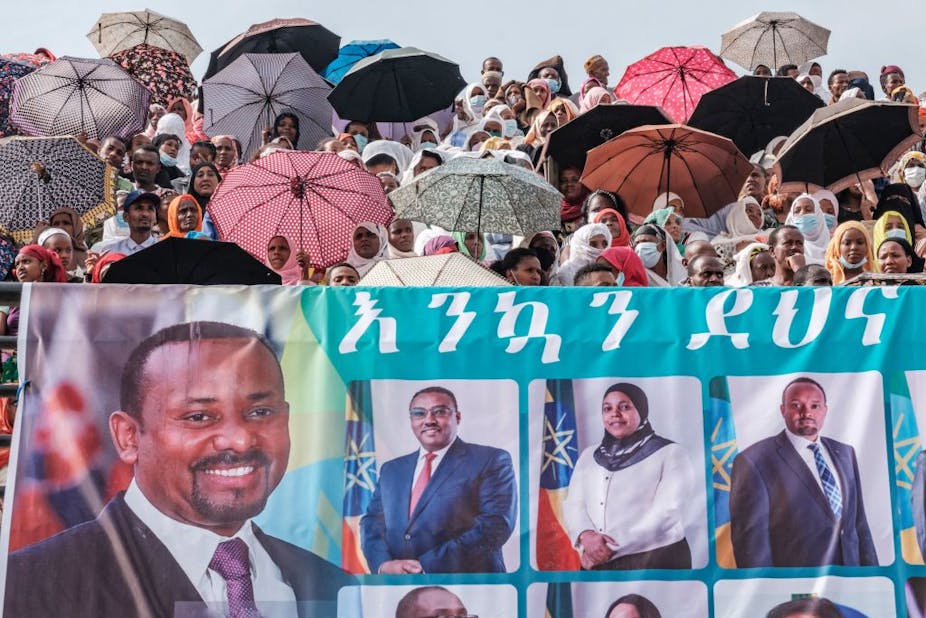 Crowd of people, some with umbrellas, and a large banner in the foreground depicting portraits of people