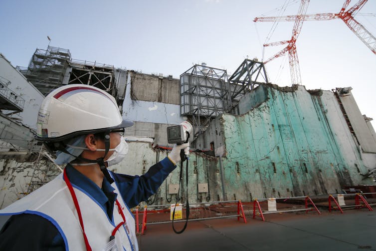 A man in a mask and hard hat holds a device up to a ruined building.