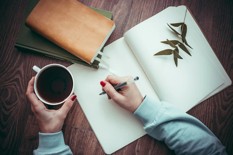 The image shows an open notebook, and a person holding a cup of coffee in one hand and pen in the other, ready to start writing.