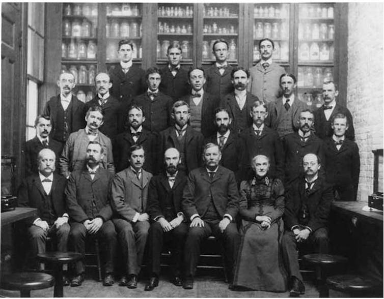 A group of people stand and sit pictured for a class-like photo. Black and white group of 20+ people mostly men