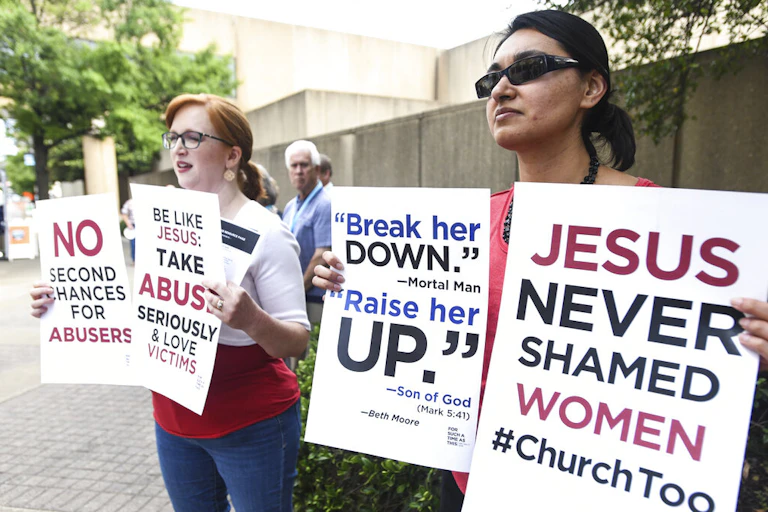 Two young women holding signs with slogans demanding action on church leaders' abuse of women and an end to shaming women victims of sexual abuse, outside a Southern Baptist Convention meeting in 2019.