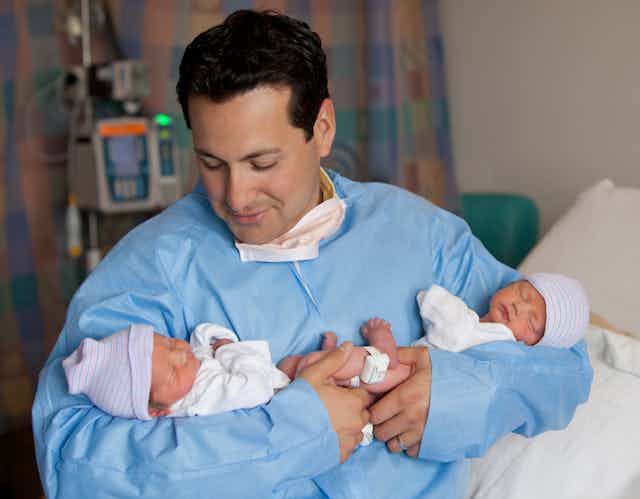 Hispanic man in a blue paper medical gown holding twin newborn babies.