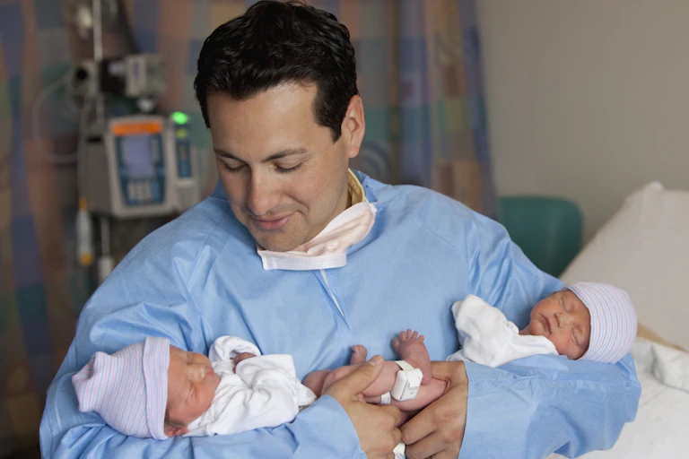 Hispanic man in a blue paper medical gown holding twin newborn babies.