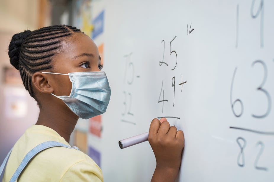 A young school girl wearing a face mask doing a maths calculation on a writing board.