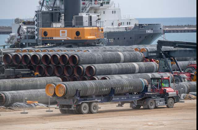 A truck drives two large pipes for the Nord Stream 2 Baltic Sea gas pipeline past several rows of similar pipes. In the background is a large ship and a crane to load the pipes.
