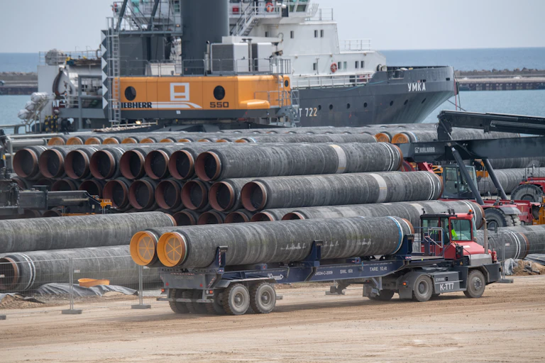 A truck drives two large pipes for the Nord Stream 2 Baltic Sea gas pipeline past several rows of similar pipes. In the background is a large ship and a crane to load the pipes.