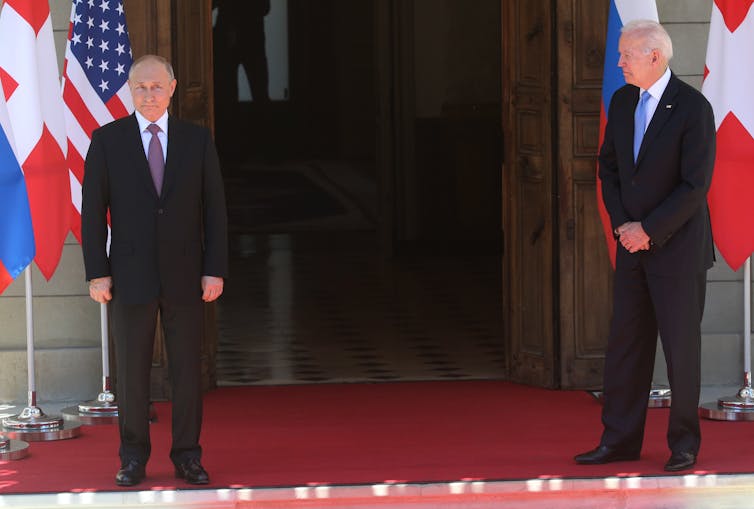 Vladimir Putin (left) and Joe Biden (right) stand in front of a door. Putin is facing the camera with a neutral expression. Biden is turned slightly to the left to look at Putin.